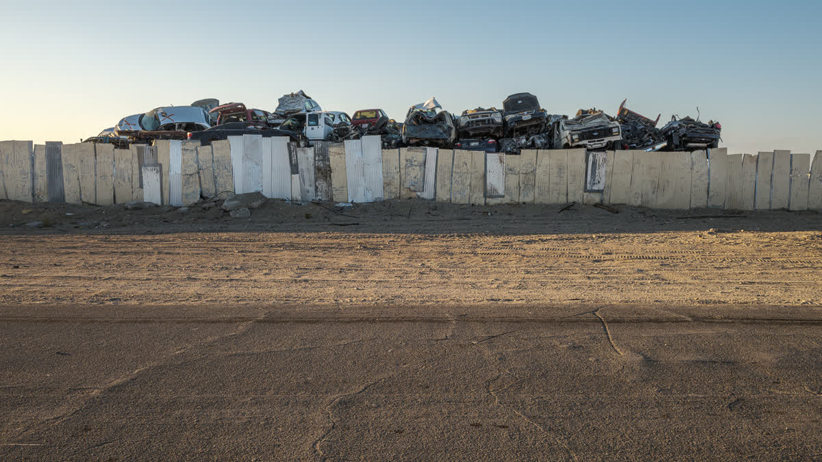 A pile of wrecked cars showing over the top of a makeshift board fence, with sand and paved road in foreground.