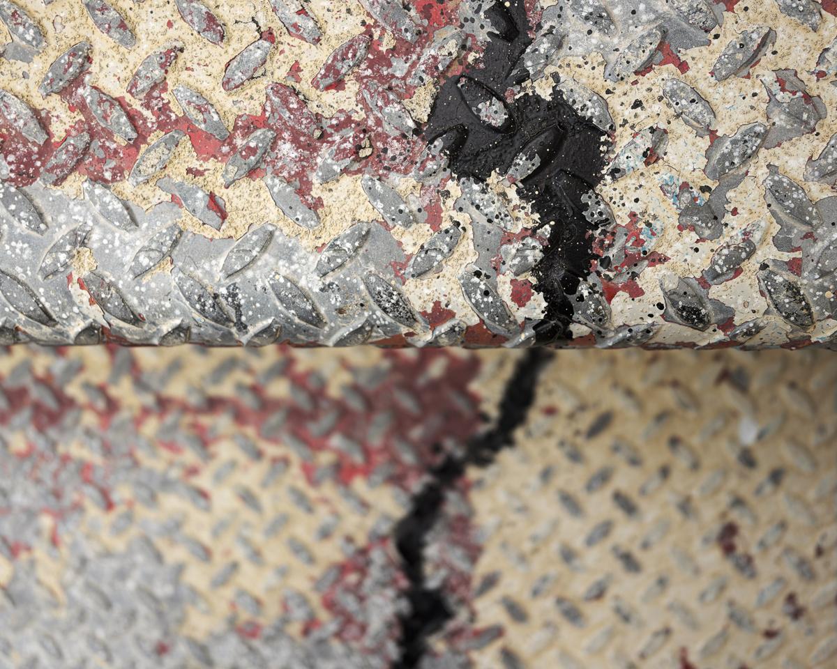 Two steps of a corrugated metal staircase with dilapidated paint, seen from directly above. The bottom step is out of focus. A new-looking black streak of paint runs across both steps, seemingly without interruption.
