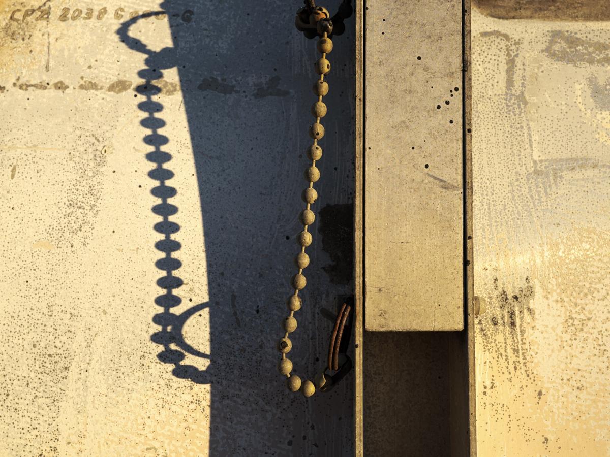 A metal partition seen edge-on, with attached chain, casts a strong shadow on a corroded metal background. 