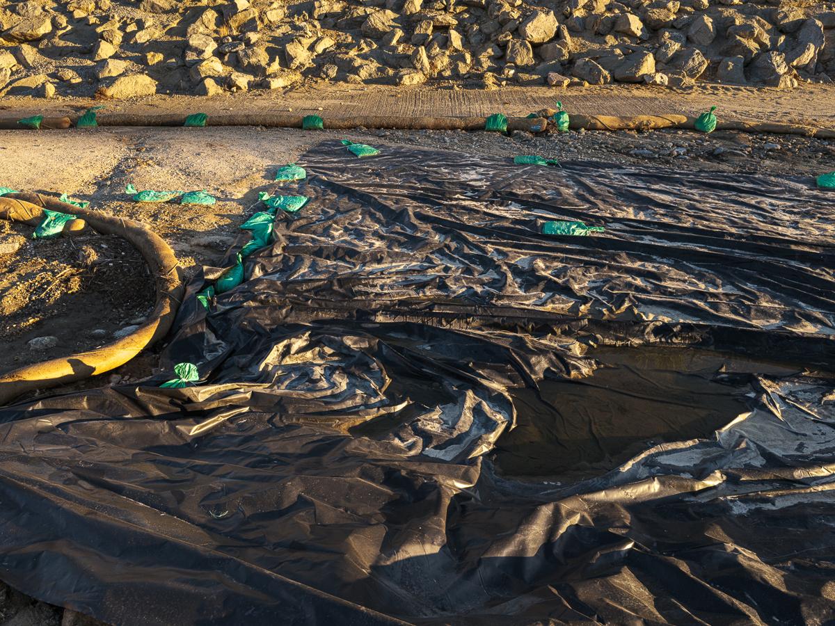 A crumpled tarp weighted down by bright green sandbags with bank of rocks in background.