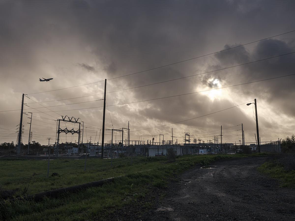 Plane ascends into cloudy sky with sunburst, over power substation with mud and grass in foreground.