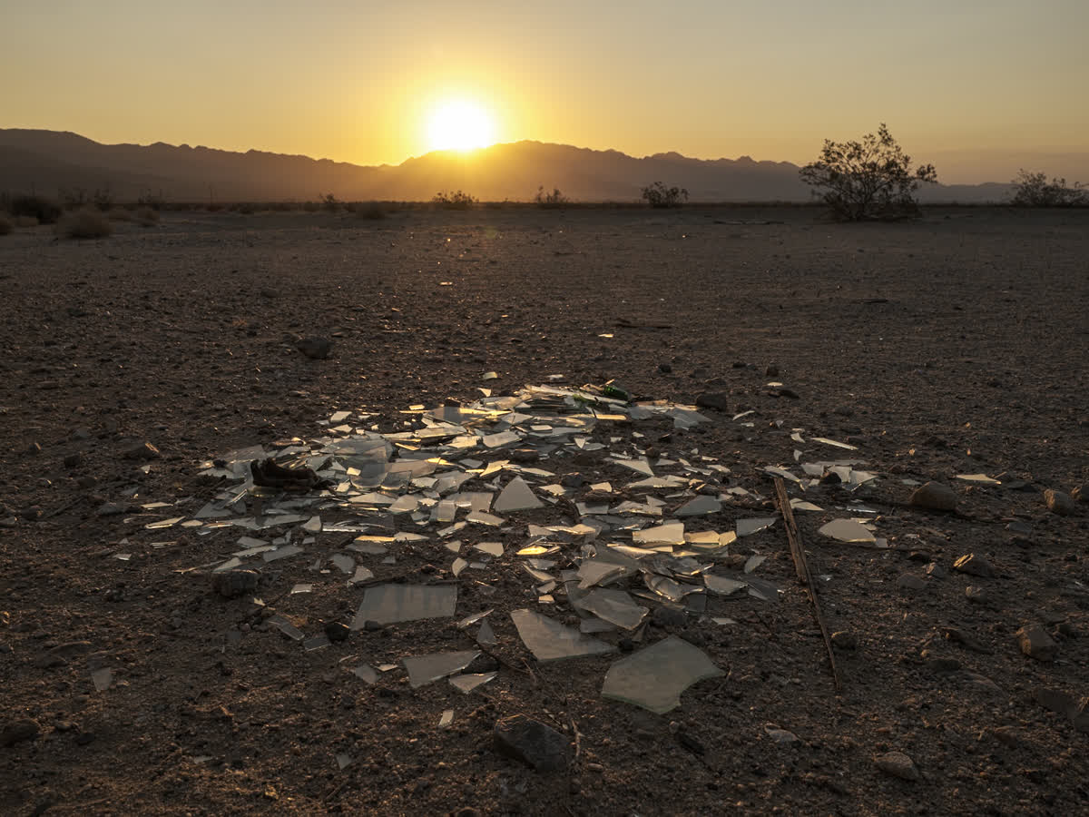 A diamond-shaped configuration of broken glass lying in desert sand with sun rising over mountains in background.