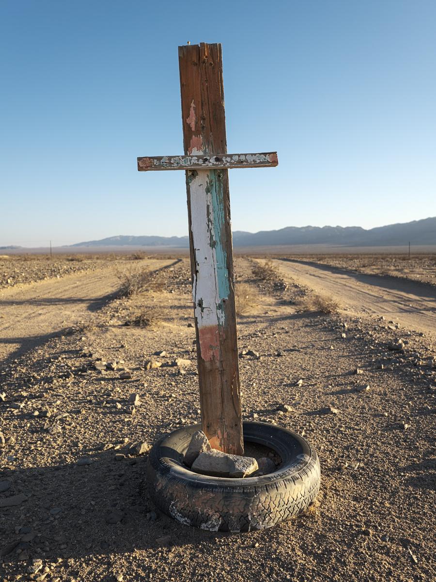 Crude wooden cross with tire base standing at a slant between two parallel dirt roads in desert landscape.
