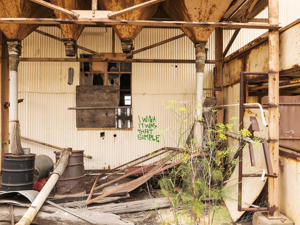 A jumble of debris, rusty pillars, broken windows, and corrugated metal with the graffito 
