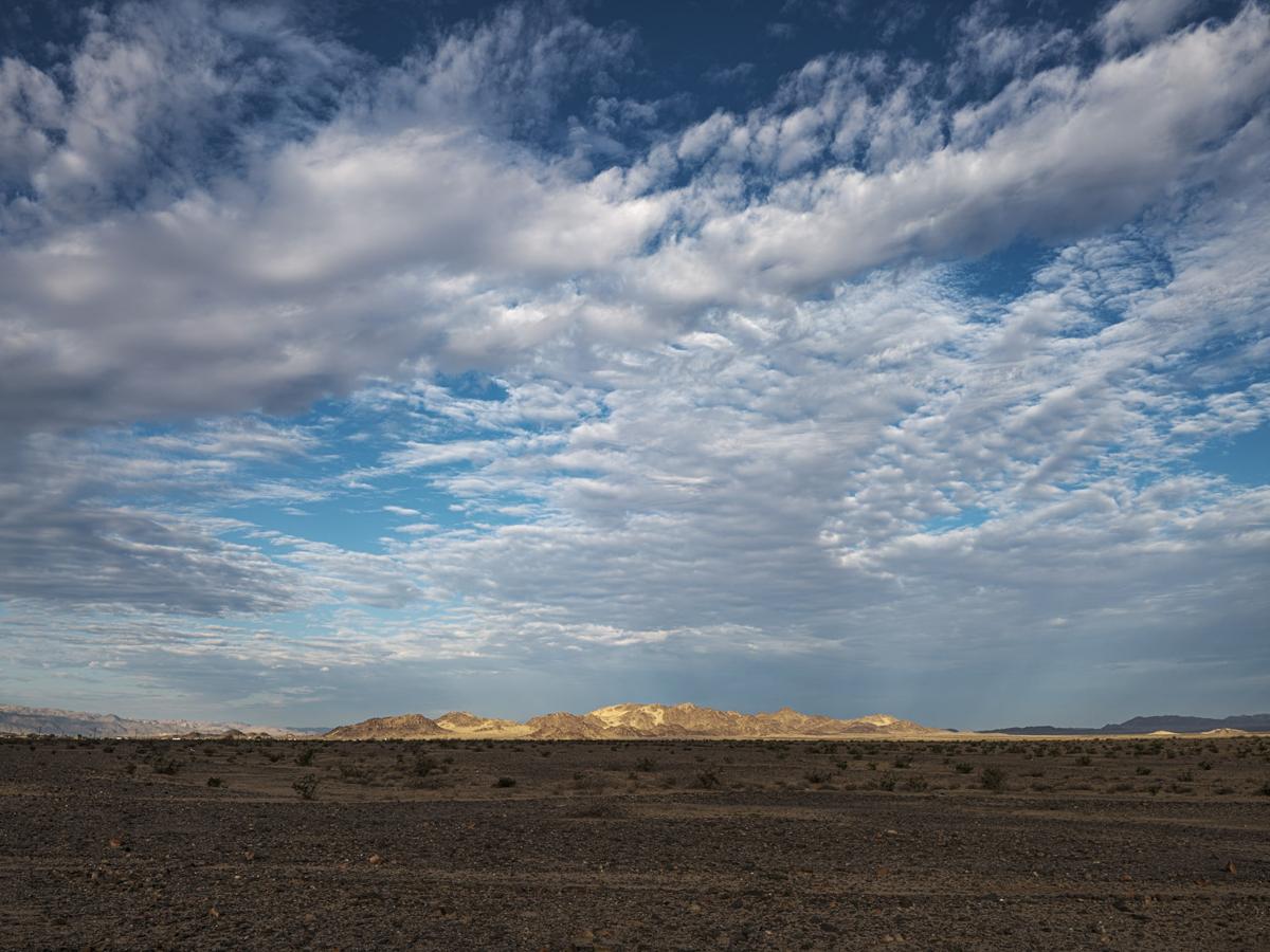 Mountain lit by sunrise with flat dark sandy desert in front and a partly cloudy sky.
