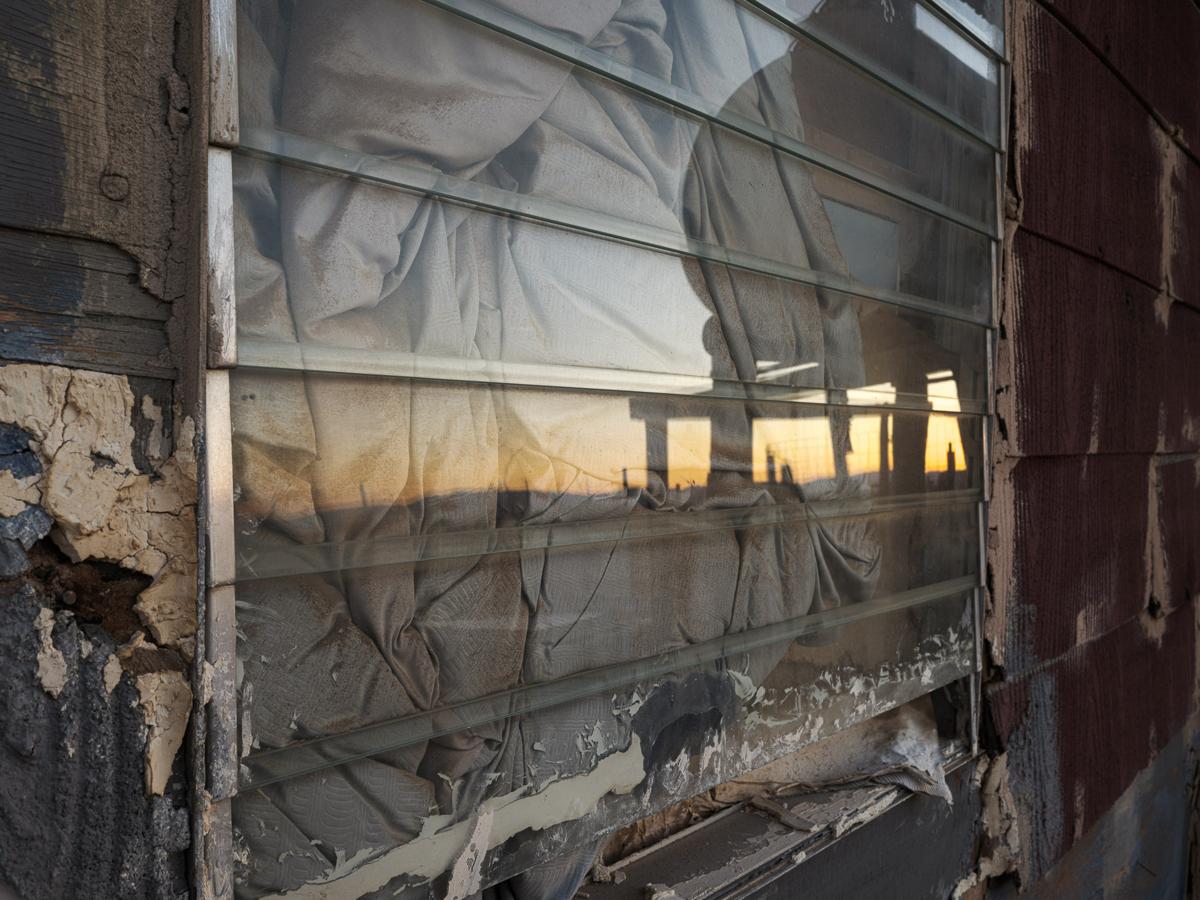 Louvred window in ruined wall with stuffing behind it, reflecting the silhouette of a water tank in dawn light.