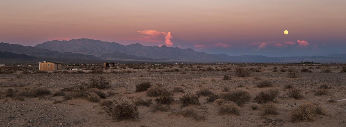 Panoramic view of full moon rising in an orange-purple sky with scattered clouds, over desert landscape with mountains in background and house in middle ground.