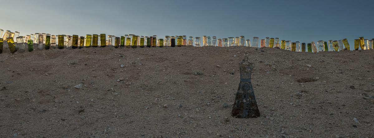 A metal dress dummy stands against a hill of sand topped by a long row of bottles lit by sunset.