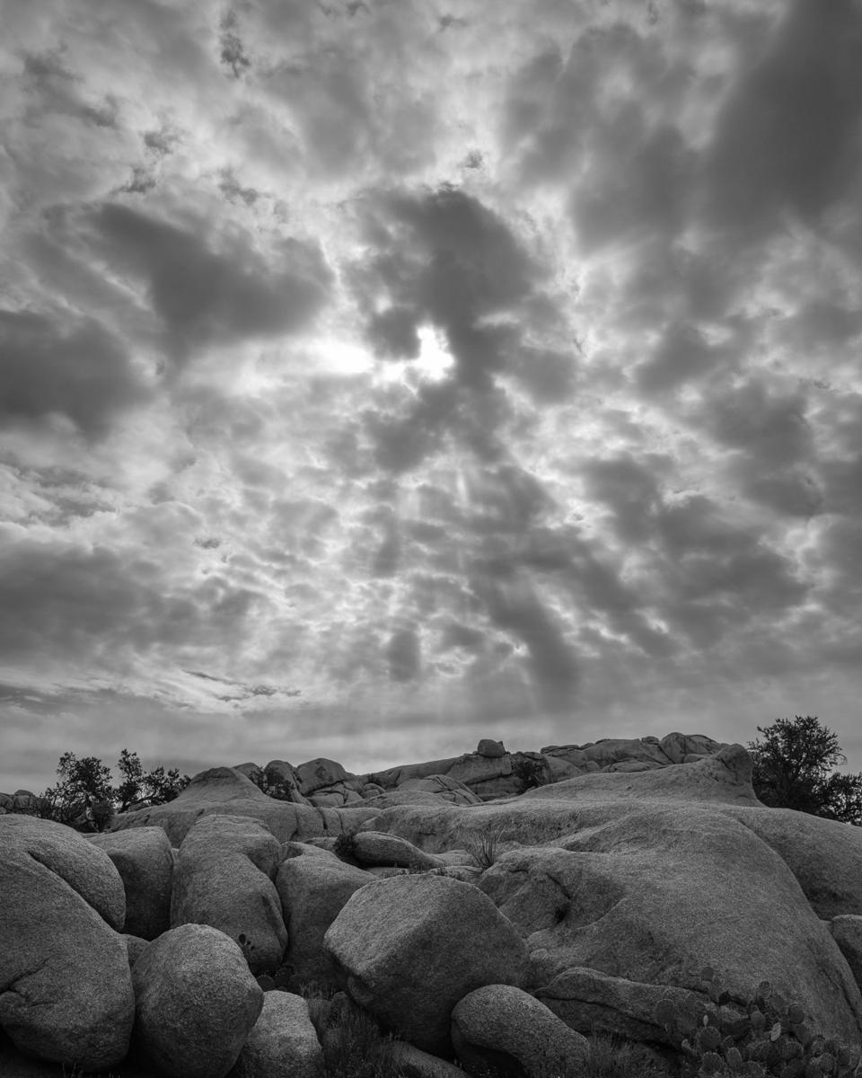Sun rays burst through altocumulus clouds with rocks in foreground.