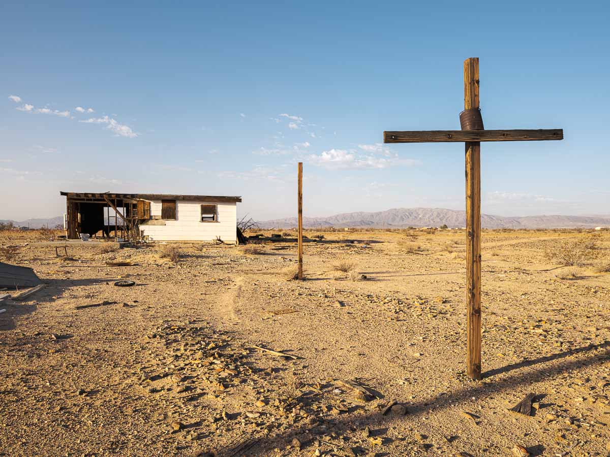 A wooden cross in front of an abandoned cabin in desert landscape.