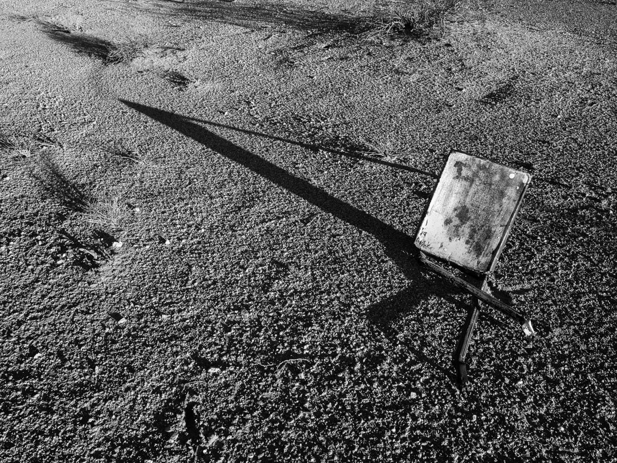 A rusty metal object casts a long and sharp triangular shadow in desert landscape.