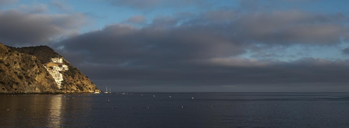 Panoramic view of ocean with headland on left. An all-white housing community is set into a cleft in the headland and spotlit by the sun.