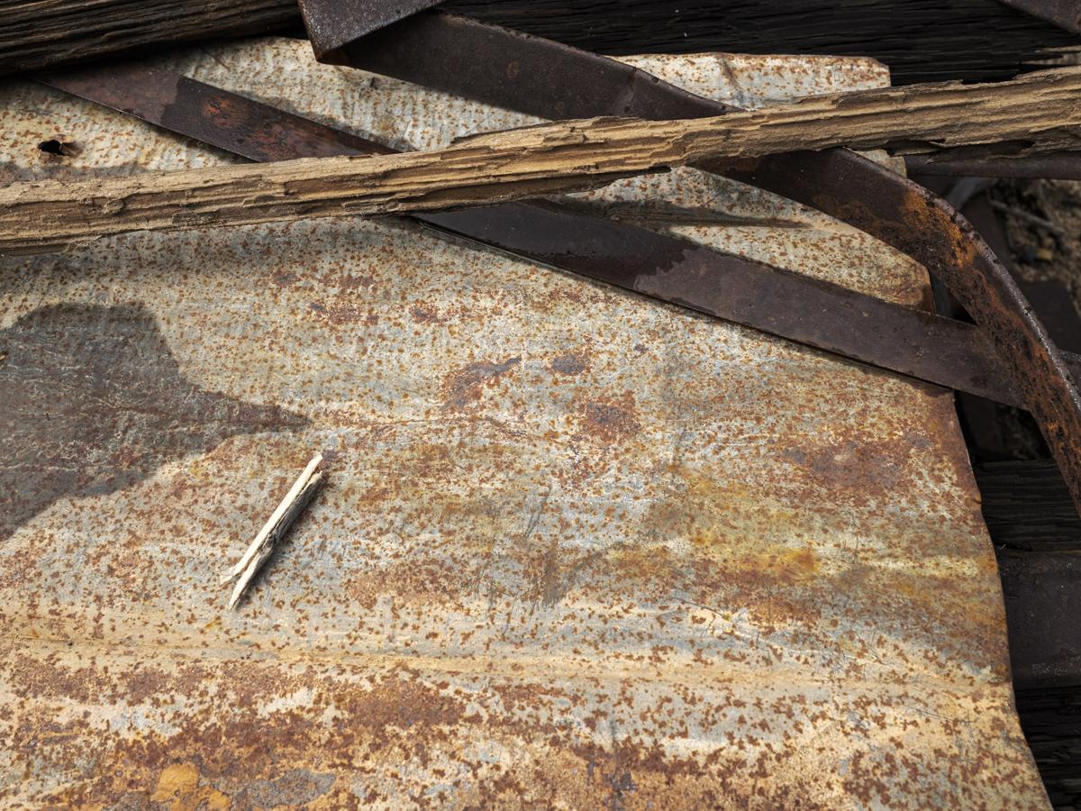 A wooden splinter rests on a sheet of partially corroded metal, with sticks and rusty struts in the background.