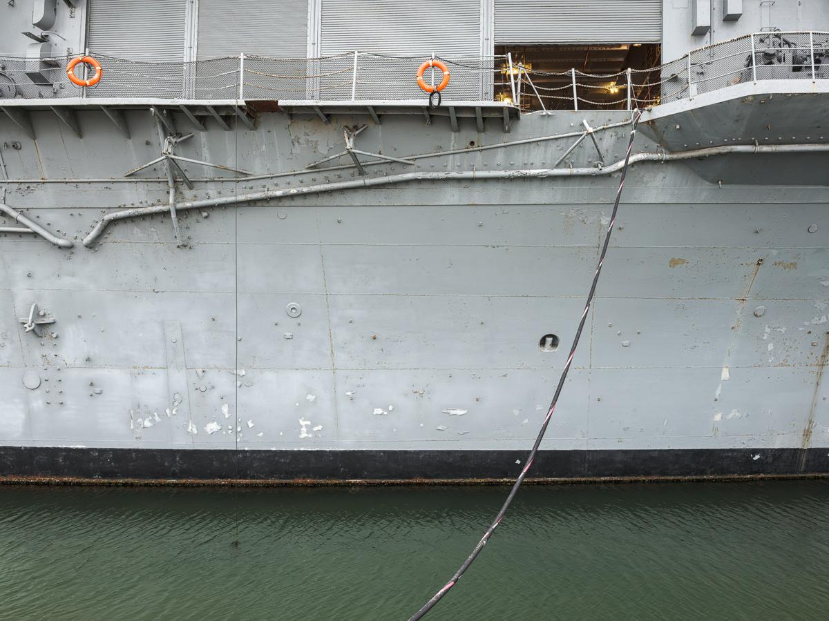 Section of ship's hull with green water below, orange life savers, and striped cord leading to dock.