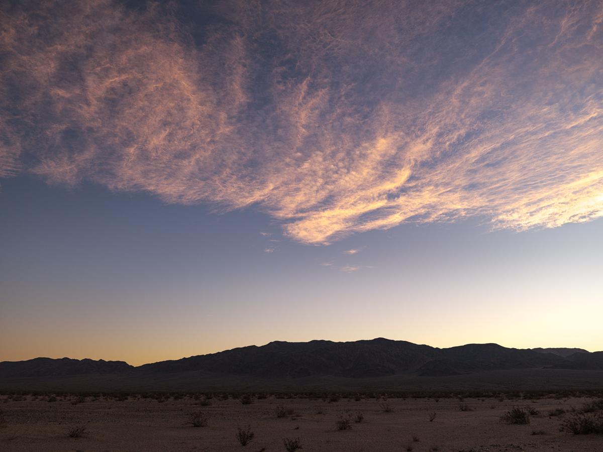 Cloud formation resembling wave lace on beach, lit by pre-sunrise light, over desert landscape with mountains.