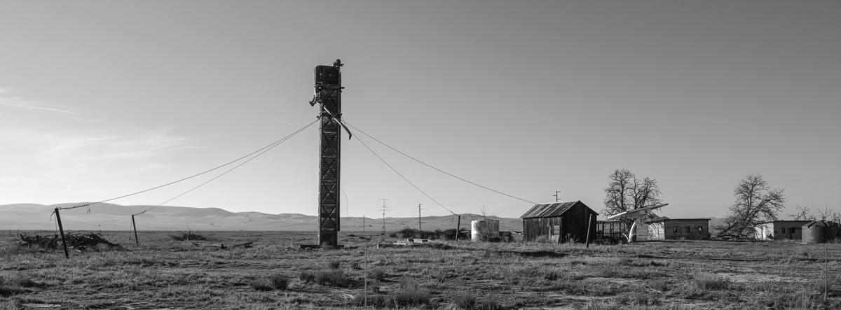 A mysterious tower is held fast by four guy ropes in desert landscape, with dilapidated huts to one side.