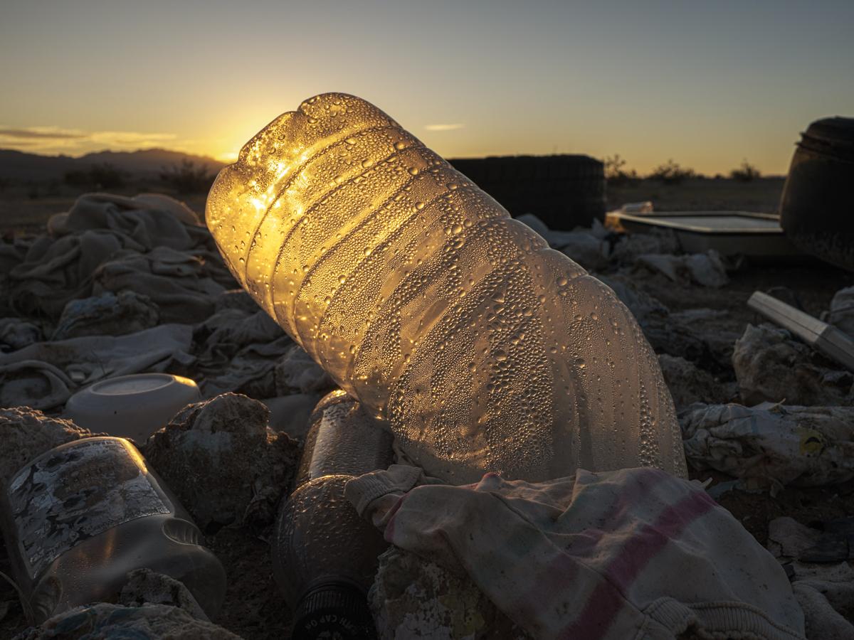 Plastic bottle covered in water droplets, lit from behind by rising sun, amid a field of trash.