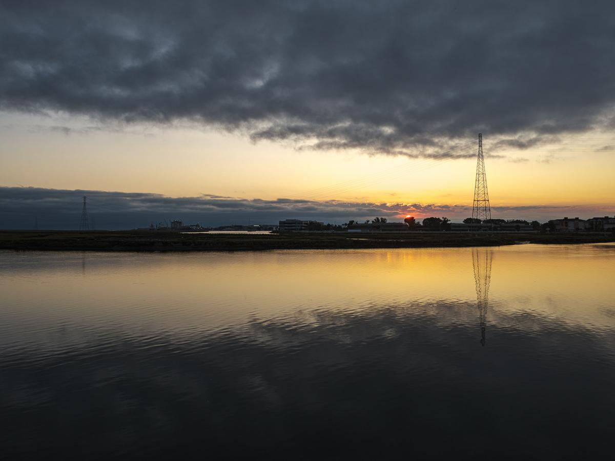 Thick cloud bank reflected in water with sun rising above shore with low buildings and power pylons.